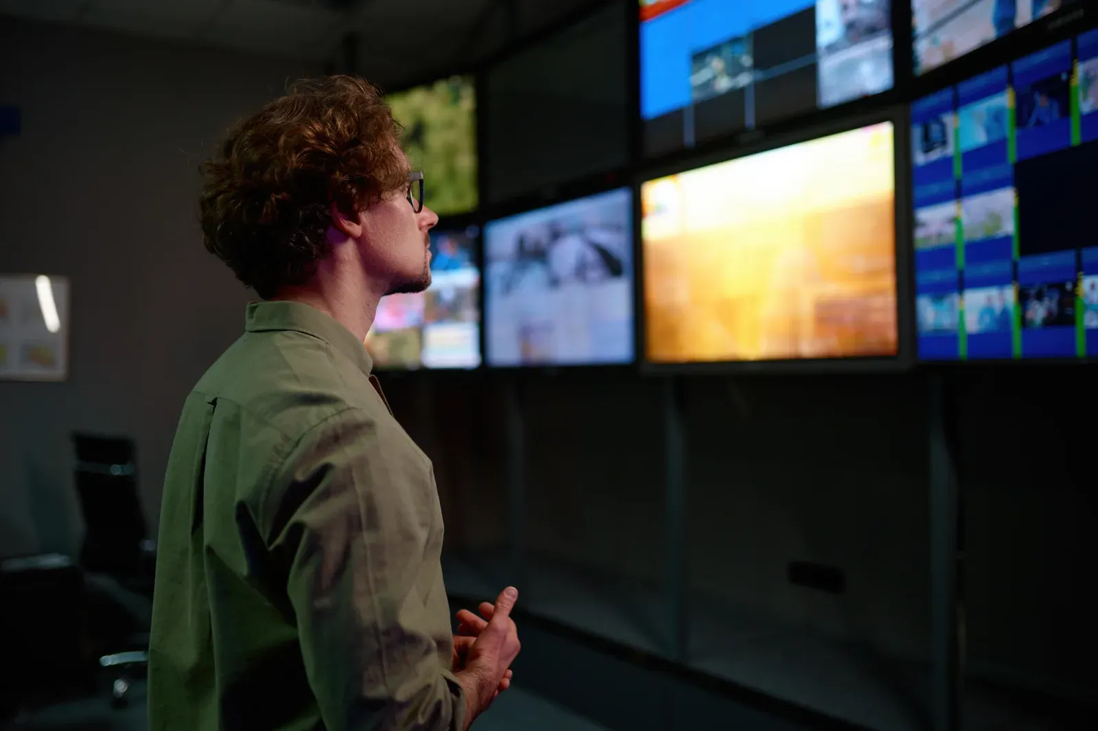 Man in profile watching a wall of TV monitors playing different content in a dark control room